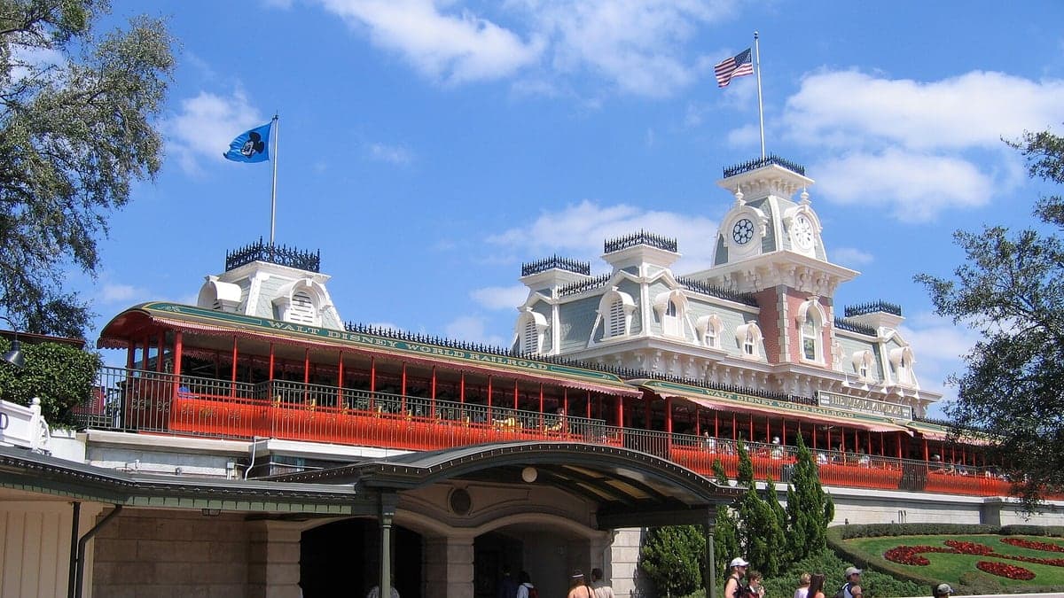 Entrada do parque temático Walt Disney World Railroad com bandeira azul com símbolo do Mickey e bandeira americana no topo do prédio com céu azul e poucas nuvens